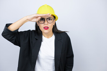 Young architect woman wearing hardhat very happy and smiling looking far away with hand over head. searching concept.