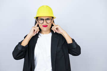 Young architect woman wearing hardhat covering ears with fingers with annoyed expression for the noise of loud music. Deaf concept.