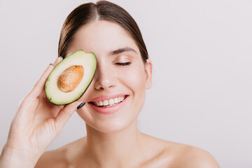 Beautiful girl without makeup posing with closed eyes on white background. Smiling model covers face with wholesome avocado