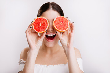 Happy girl with positive emotional facial expression covers her eyes with oranges, posing on isolated background
