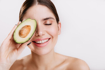 Closeup portrait of young lady covering eye with avocado. Brunette girl smiling friendly on white background