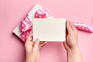 Greeting card mockup with gift box with bow in female hands on pink paper background