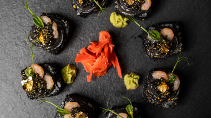 Sushi roll with nori, black rice, crab meat, cucumber, avocado, smoked salmon mousse, oar caviar, masago, shrimp cocktail and edible gold leaf with red ginger and wasabi on black table and background.