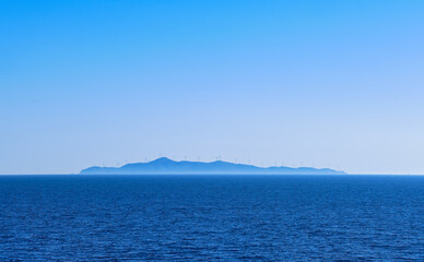 Seascape of calm azure sea, blue sky, no clouds. Distant island in haze with silhouettes of electric turbine windmills. Mediterranean sea, Greece.