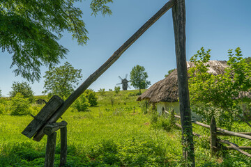 Fototapeta na wymiar Old windmill and medieval, traditional Ukrainian rural houses on a green meadow. Blue sky with white clouds.