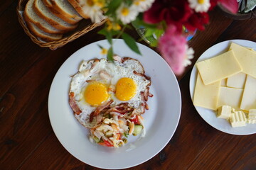 A breakfast with fried eggs, salad and cheese is served in white plates on a wooden table. The flowers are decorating the breakfast. This is a simple food of everyday people plating their meals at