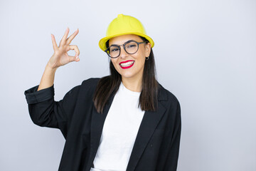 Young architect woman wearing hardhat doing ok sign with fingers and smiling, excellent symbol