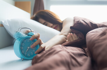 Obraz na płótnie Canvas young asian woman sleeping on bed pressing snooze button on blue alarm clock at seven o'clock morning in bed room at home, lifestyle, good morning, healthy sleep and joyful weekend concept