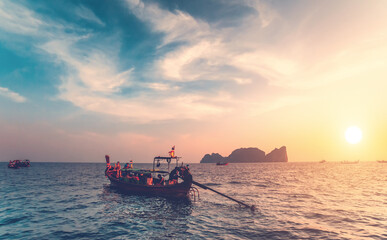 Obraz premium Thailand ocean boats cruise: people traveling on water transport at sun setting tones. Majestic sunset tourist vacation at waterfront. Mountain island silhouette at skyline under cloudy sky