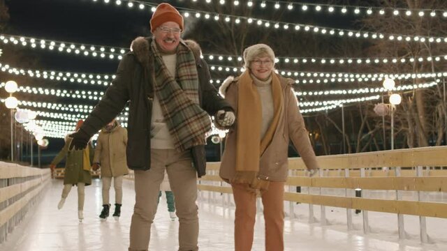 Full shot of cheerful senior couple and young family of three skating on outdoor ice rink together holding hands together in winter evening