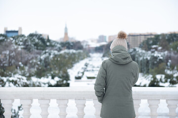Mujer joven de pie con abrigo verde y gorro de lana, dando la espalda a la cámara y mirando un parque nevado vacío - obrazy, fototapety, plakaty