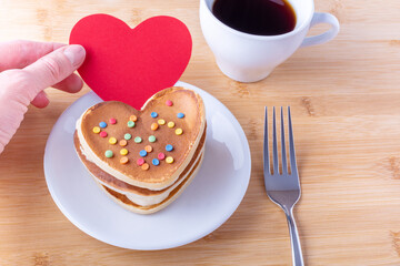 Hand holds a red blank heart shape next to homemade heart shaped pancakes with sugar decoration on a white plate, fork, mug with coffee or cocoa, close-up. Breakfast for Valentine's Day Concept