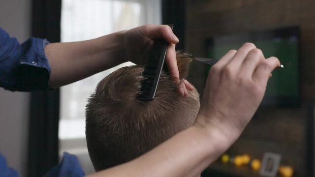 Wife cutting husband's hair at home in front of TV with scissors and holding comb in other hand.
