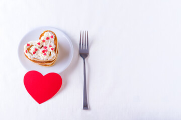 Homemade heart shaped pancakes decorated with cream and sugar candy on a white plate, fork and red blank heart shape on a white background, top view, copy space. Postcard for Valentine's Day