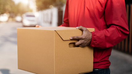 Cropped shot of a delivery man in red uniform holds postal parcel stands on street looks at...