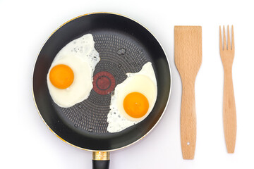 Fried eggs in a frying pan with a wooden fork and spatula isolated on white.