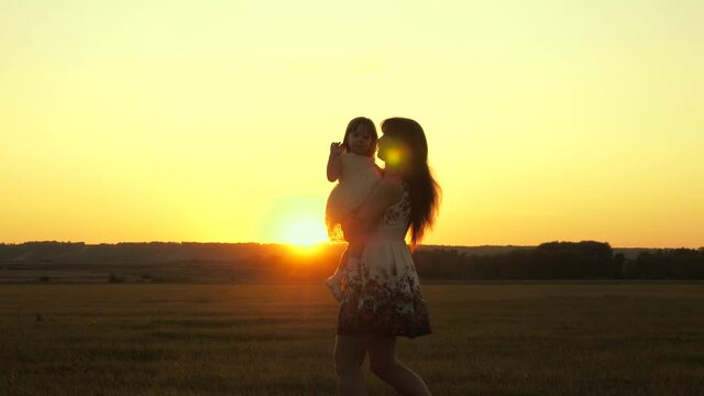 Mom is carrying her beloved healthy daughter in her arms, a walk in park in summer. Child hugs and kisses mommy in sun. Mother plays with her baby. A child with parent walks at sunset. Happy family