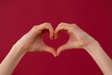 Obraz premium Female hands showing heart sign on red background in studio. Fingers illustrating gesture of love on burgundy backdrop. Valentine theme expressed in symbol.