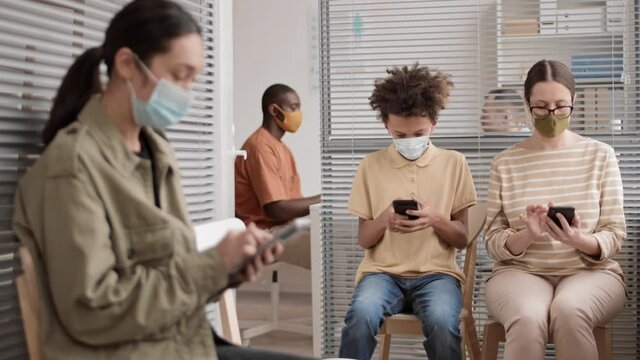 Medium long steadicam of two Caucasian women and African school-aged boy wearing masks, sitting in hall of medical office, using smartphones, waiting their turn, man talking to doctor on background