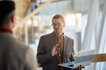 young businessman with glasses, piercing and earrings holding laptop  talking to a colleague