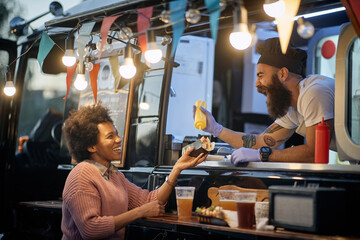 Fototapeta na wymiar young beardy caucasian employee in fast food laughing, adding a mustard in a sandwich to a female afro-american customer