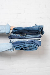 partial view of woman holding stack of blue denim clothes near white brick wall