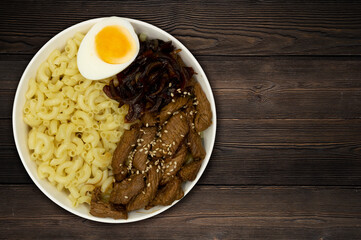 Meat with caramelized onions, egg and pasta on a wooden background