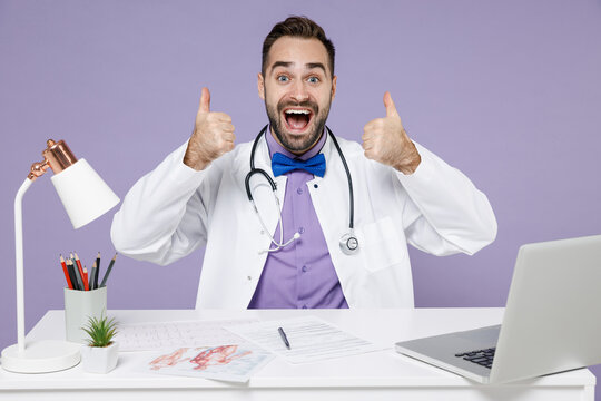 Smiling Male Happy Doctor Man In White Medical Gown Suit Sit At Desk Work On Computer Documents In Clinic Office Show Thumb Up Gesture Isolated On Violet Background Studio Portrait Healthcare Concept.