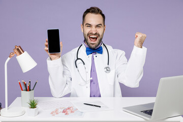 Male lucky doctor man in white medical suit sit at desk work in clinic office hold mobile phone blank screen workspace area do winner gesture clench fist isolated on violet background studio portrait.