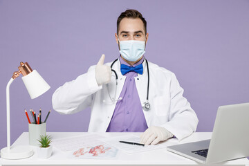 Male doctor man in white medical suit sit at desk work in clinic office in sterile face mask, from coronavirus virus covid-19, latex gloves show thumb up isolated on violet background studio portrait