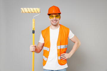 Young employee man in orange vest protective hardhat holding paint roller stand akimbo arm on waist isolated on grey background studio. Instruments for renovation apartment room. Repair home concept