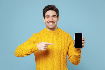 Young smiling happy student man 20s in knited yellow sweater hold in hand using point index finger on mobile cell phone with blank screen workspace area isolated on blue background studio portrait. - Powered by Adobe