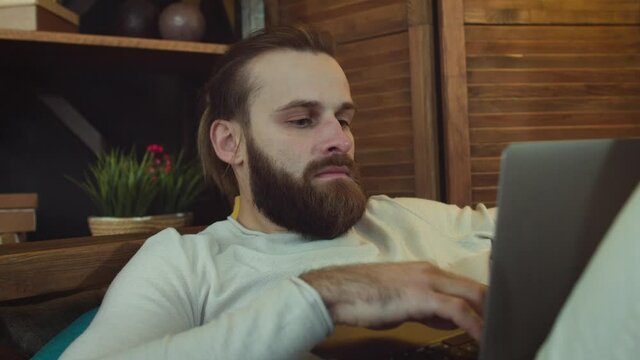 Caucasian Man with Laptop. He is Working at Home oo a sofa. Typing and Smiling 
