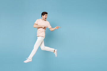 Fototapeta na wymiar Full length of young surprised cheerful smiling student man 20s in beige t-shirt white pants point index finger aside on copy space workspace area jump run isolated on blue background studio portrait.