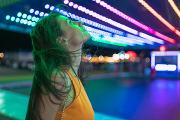 Side shot of thoughtful girl with blown hair looking up at the lights in Amusement park at the night