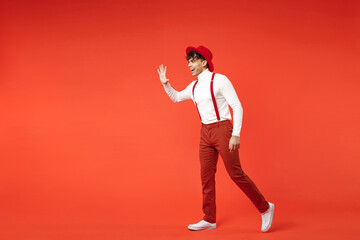 Full length of young spanish latinos attractive handsome smiling stylish fashionable man 20s in hat white shirt trousers with suspenders walk going isolated on bright red background studio portrait.