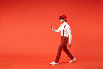 Fototapeta na wymiar Full length side view of young spanish latinos smiling fashionable man 20s wearing hat white shirt trousers, suspenders walk go hold mobile cell phone isolated on red color background studio portrait.