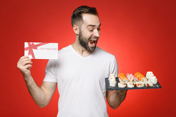 Surprised young bearded man 20s in white t-shirt hold in hand gift certificate makizushi sushi roll served on black plate traditional japanese food isolated on red color background studio portrait.