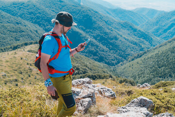Young man checking GPS on his smartphone while on a hiking trip near mountain edge with green hills