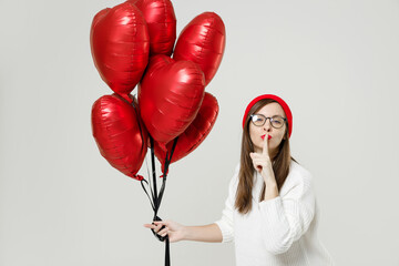 Secret young woman in sweater red hat glasses say hush be quiet with finger on lips shhh gesture celebrating birthday holiday hold bunch heart air inflated helium balloon isolated on white background.