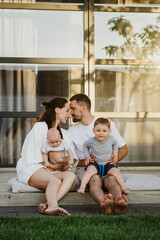 A young beautiful family with two little sons hug and kiss in the yard of their own house. Young mom and dad with two young sons hug and kiss in the yard of their own house.