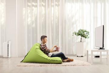 Teenager sitting on a bean bag armchair with a box of popcorn and watching tv in a minimal modern living room