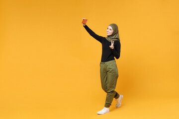 Fototapeta na wymiar Full length of funny young arabian muslim woman in hijab black green clothes doing selfie shot on mobile phone showing victory sign isolated on yellow background. People religious lifestyle concept.