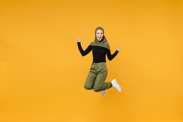 Fototapeta na wymiar Full length happy young arabian muslim woman in hijab black green clothes jumping clenching fists like winner say yes isolated on yellow background studio portrait. People religious lifestyle concept.