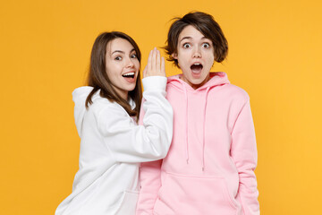 Shocked funny two young women friends 20s in casual white pink hoodies whispering gossip and tells secret behind her hand, sharing news isolated on bright yellow color background studio portrait.
