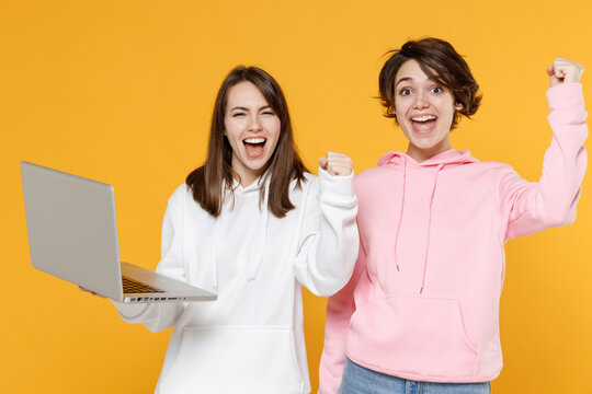 Joyful two young women friends 20s wearing casual white pink hoodies standing working on laptop pc computer doing winner gesture say yes isolated on bright yellow color background studio portrait.