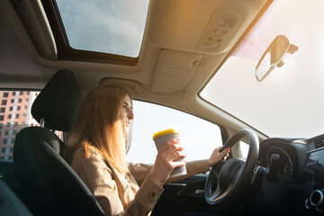 Young woman focused on driving holds a cup of coffee or tea . Sun rays on the windshield. Driving concept