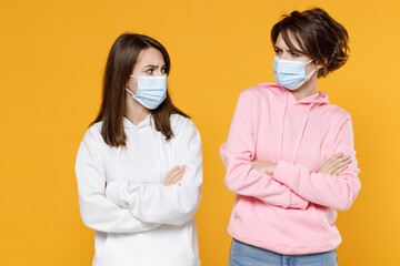 Displeased two young women friends 20s in casual hoodies sterile face mask safe from coronavirus virus covid-19 hold hands crossed looking at each other isolated on yellow background studio portrait.