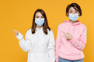 Shocked two young women friends 20s in basic white pink hoodies sterile face mask safe from coronavirus virus covid-19 pointing index fingers aside isolated on yellow color background studio portrait.