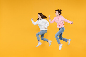 Fototapeta na wymiar Full length side view of cheerful two young women friends 20s wearing basic white pink hoodies jumping running clenching fists like winner isolated on bright yellow color background studio portrait.